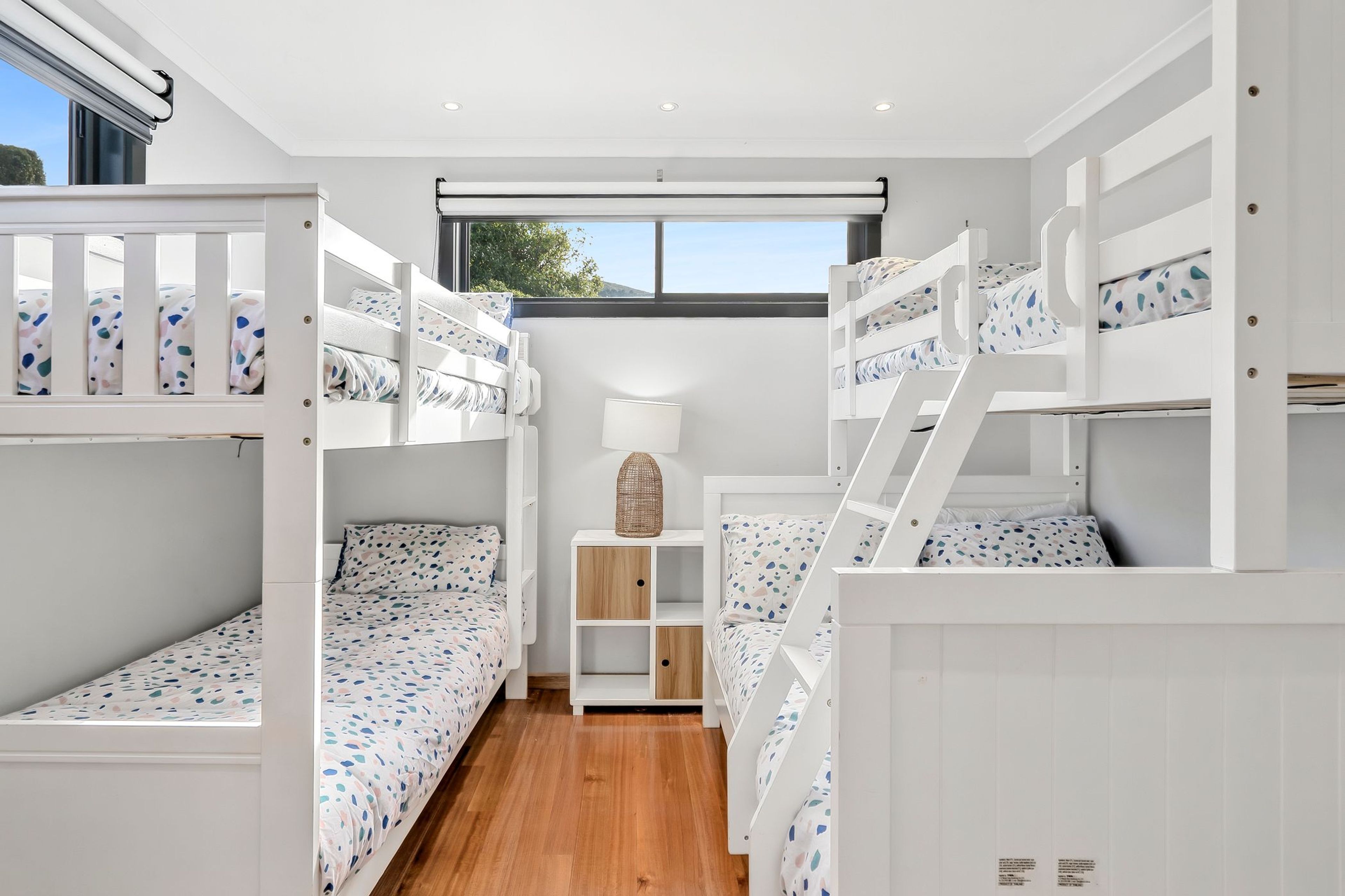 A children's bunk room with white bunk beds, a wooden floor, and a window with a view of trees.