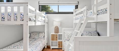 A children's bunk room with white bunk beds, a wooden floor, and a window with a view of trees.