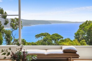 Ein Zimmer mit großem Fenster und Blick auf die Küste, eine Holzbank und ein Buch.