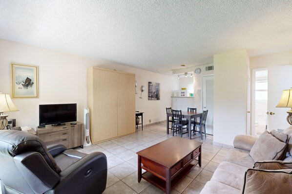 A living room with a sofa, a coffee table, a TV, and a dining area with chairs and a table. - One block from Pompano Beach - condo w/ shared heated pool & sundeck (Pompano Beach)