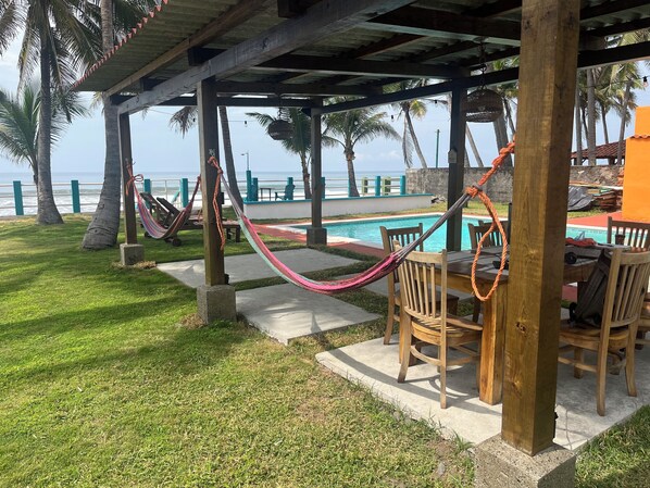 A covered outdoor seating area with a pool, wooden chairs, and a hammock.