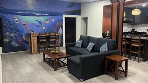 Een moderne woonkamer met een groot muurdecoratie van een onderwaterlandschap, een zwarte bank met gekleurde kussens, houten bijzettafels en een keukenhoek met houten stoelen.