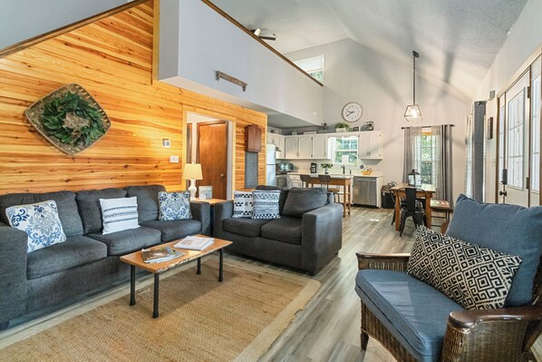 A spacious living area with two sofas, a coffee table, and a dining table with chairs. A kitchen is visible in the background with cabinets and a window. - Welcome to “Stealing Time” – Your Cozy Getaway (Burnside)
