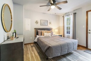 A bedroom with a bed, a ceiling fan, a window with curtains, a door, and a small table with a lamp. - Welcome to “Stealing Time” – Your Cozy Getaway (Burnside)
