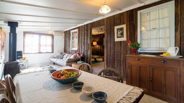 A cozy living room with a wooden wall, a dining table with a bowl of fruit, and a fireplace. - Cox's Cottage in the Hawkesbury Valley (Pitt Town)