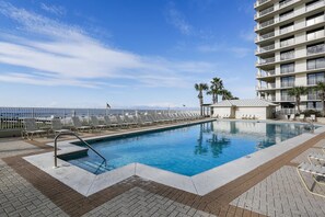 Área da piscina com vista clara para o oceano, um edifício com varandas e um espaço de estar coberto.