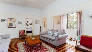A living room with a grey sofa, a wooden coffee table, a red armchair, and a red and white patterned rug. There are framed pictures on the wall, a window with curtains, and a view of trees outside. - Daves Place, 27 Rigney St - Holiday house with Wifi, Dog Friendly, Aircon & Boat Parking (Shoal Bay)