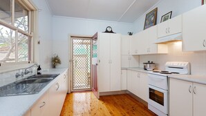 A compact kitchen with white cabinets, a wooden floor, a sink, a stove, and a clock on the wall. - Daves Place, 27 Rigney St - Holiday house with Wifi, Dog Friendly, Aircon & Boat Parking (Shoal Bay)