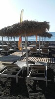 Una playa con sombrillas de paja, sillas reclinables blancas y una terraza de madera.