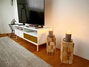 A modern living room with a white entertainment unit, a flat-screen TV, a small side table, and two candles on wooden stands.
