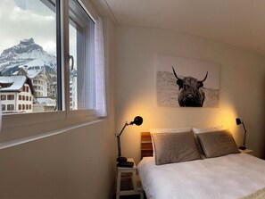 A bedroom with a bed, two bedside lamps, a window with a view of buildings and mountains, and a bull painting on the wall. - SnowKaya Appartement/Fewo 5-Bettwohnung by Interhome (Engelberg)