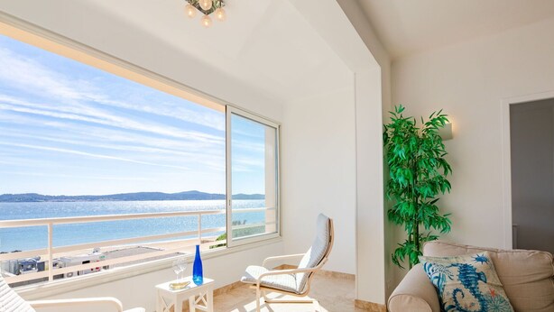 A spacious balcony with a view of the sea, a white chair, a small table with a blue vase, and a beige sofa with a decorative pillow.