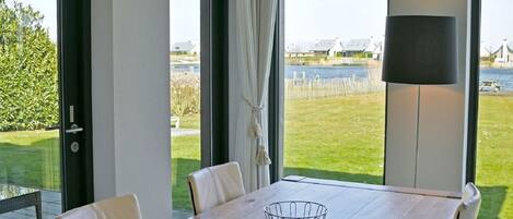A dining area with a wooden table, white chairs, and a metal bowl centerpiece. Large windows offer a view of a grassy area and a body of water.