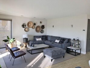 A modern living room with a grey sectional sofa, a coffee table, and a grey ottoman. There are two black chairs, a potted plant, and wall art.