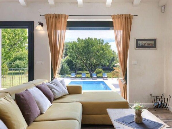 A modern living room with a beige sofa, a small coffee table, and a view of a swimming pool and outdoor seating area.
