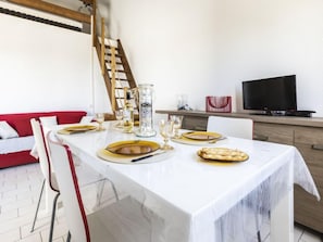 Ein Esstisch ist mit Tellern, Besteck und Gläsern gedeckt, im Hintergrund steht ein Fernseher auf einem Schrank und ein rotes Sofa.