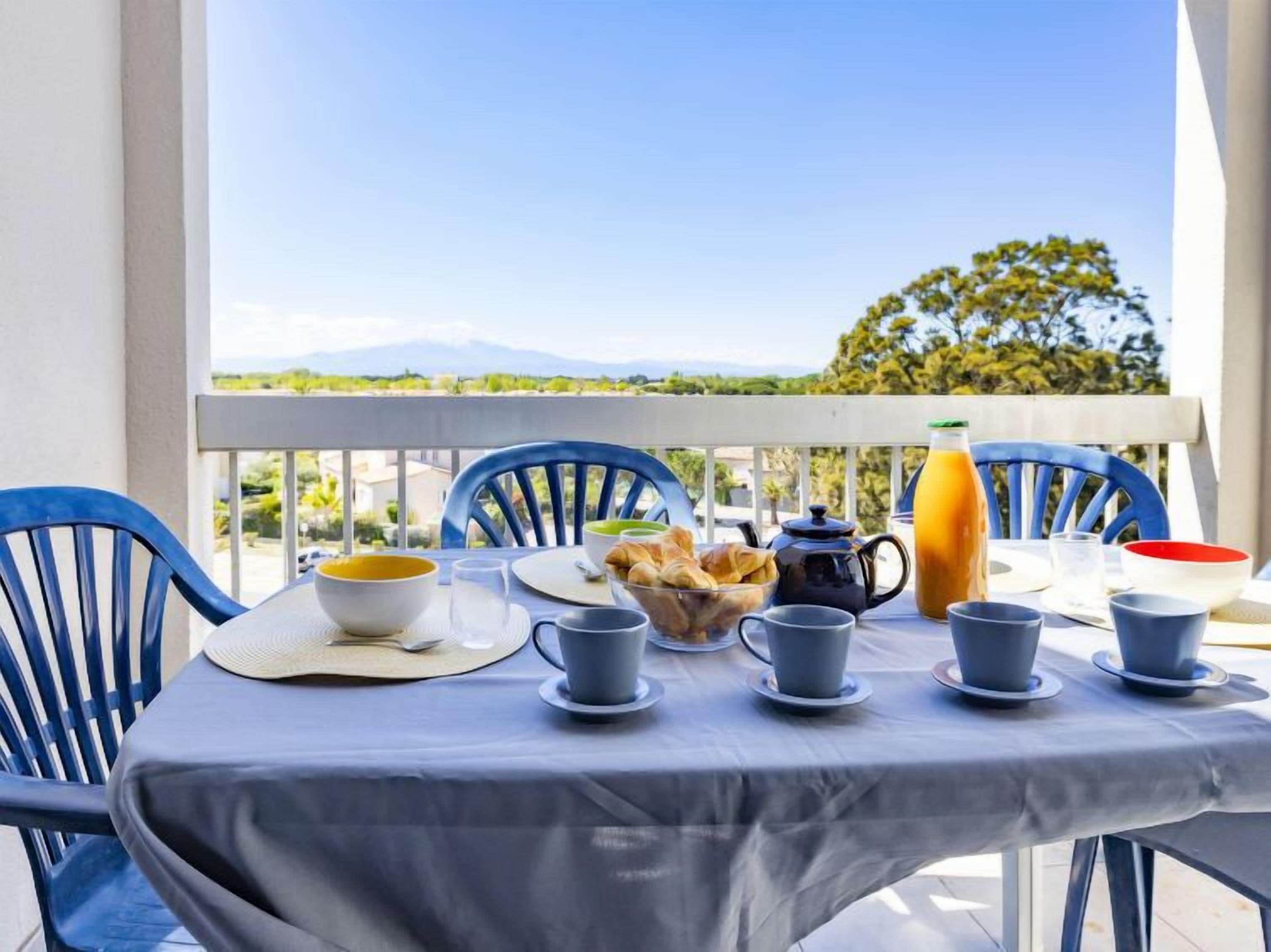 Une table de balcon dressée avec des éléments pour le petit-déjeuner, notamment des croissants, une théière et une bouteille de jus d’orange.