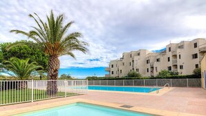 A swimming pool area with a palm tree, a white fence, and a building with multiple balconies in the background. - Les Frégates by Interhome (Saint Cyprien)