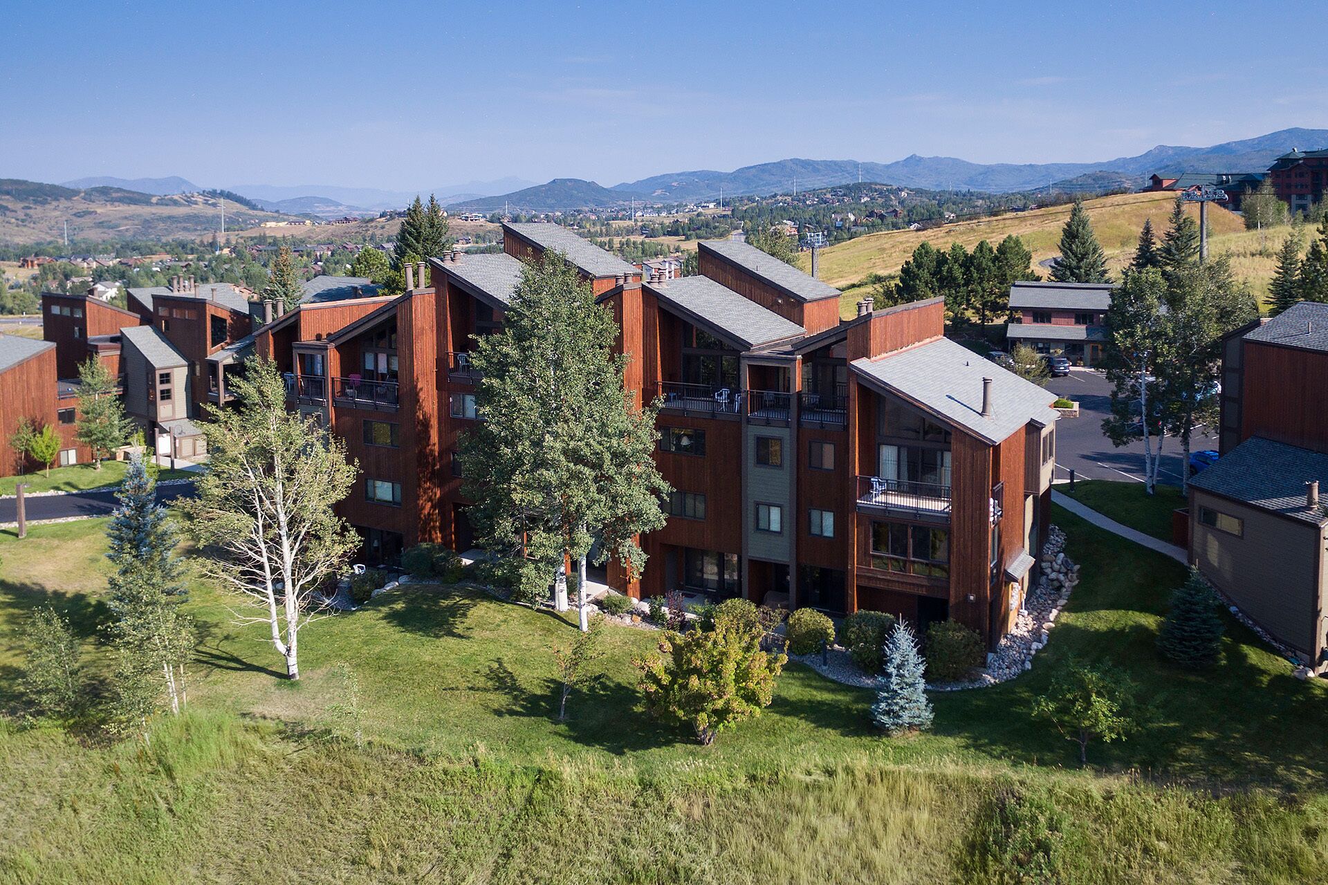 Un immeuble moderne à plusieurs étages, doté de balcons, entouré de verdure et se dressant sur un fond de montagnes.