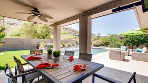 Um pátio externo com mesa de jantar posta para quatro pessoas, rodeada por cadeiras, um ventilador de teto e vista para uma piscina e vegetação.