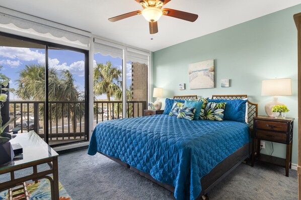 A bedroom with a large bed, a ceiling fan, a view of palm trees, and a balcony.