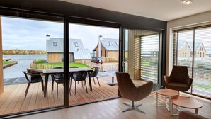 A modern interior with a large sliding glass door leading to an outdoor deck. Inside, there is a wooden table with chairs and a modern chair. - Oesterdam Resort by Interhome (Tholen)