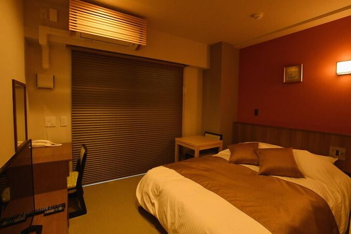 A hotel room with a bed, a desk with a telephone, a chair, a small table, and a window with blinds.