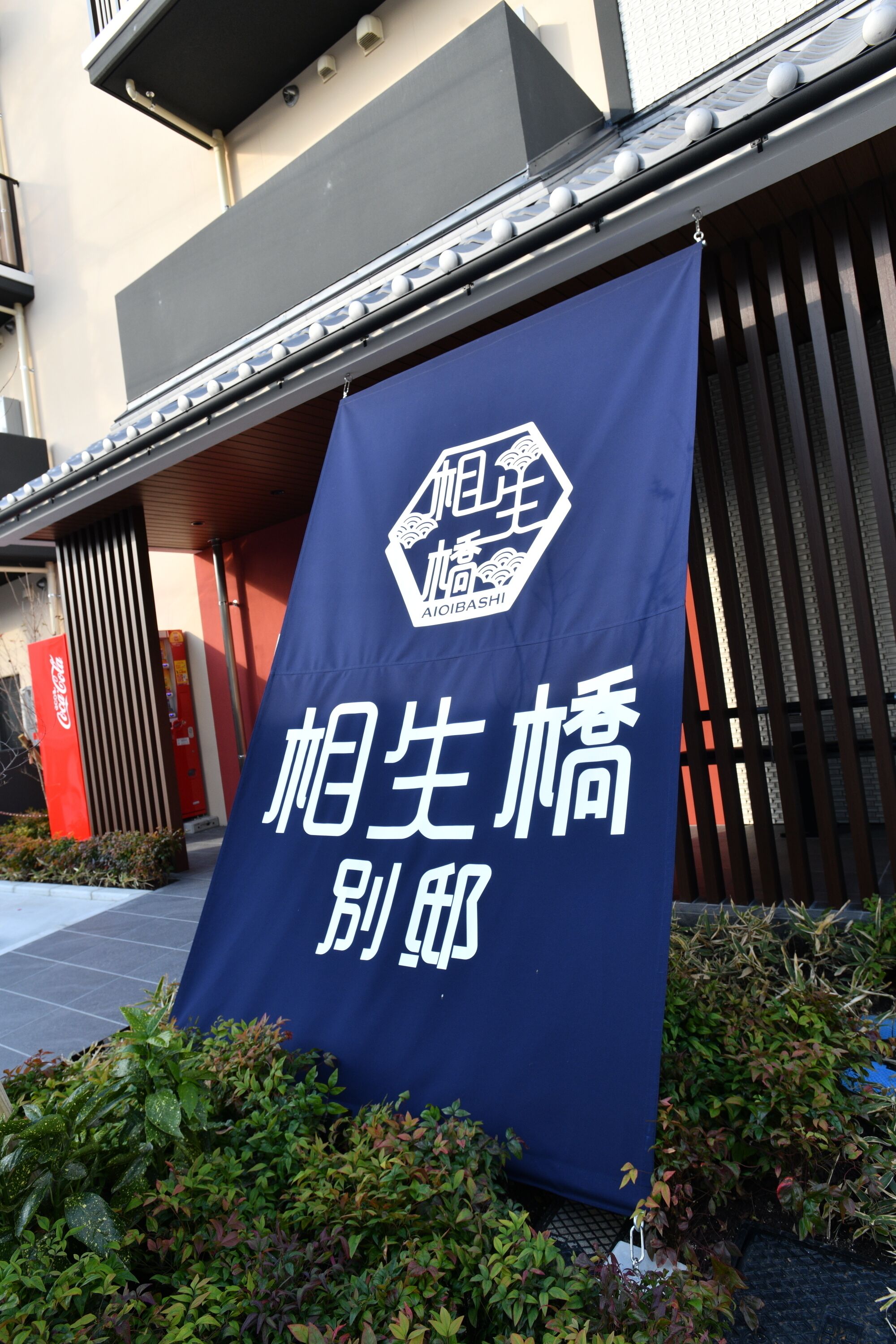 A banner with a logo and Japanese text, hanging outside a building with a sloped roof and metal railings.
