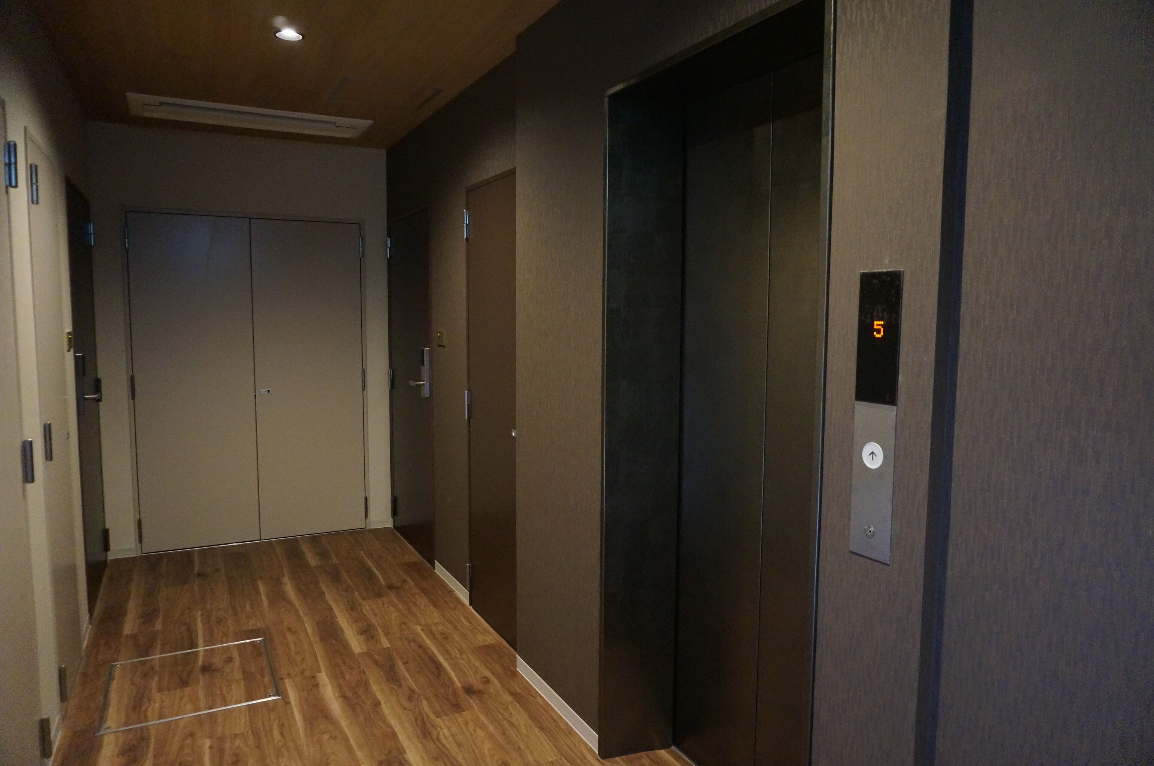 A hallway with wooden flooring, a set of double doors, and an elevator with a digital display showing the number 5.