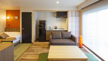 A compact hotel room with a bed, a sofa, a small dining area, and a window with blinds.