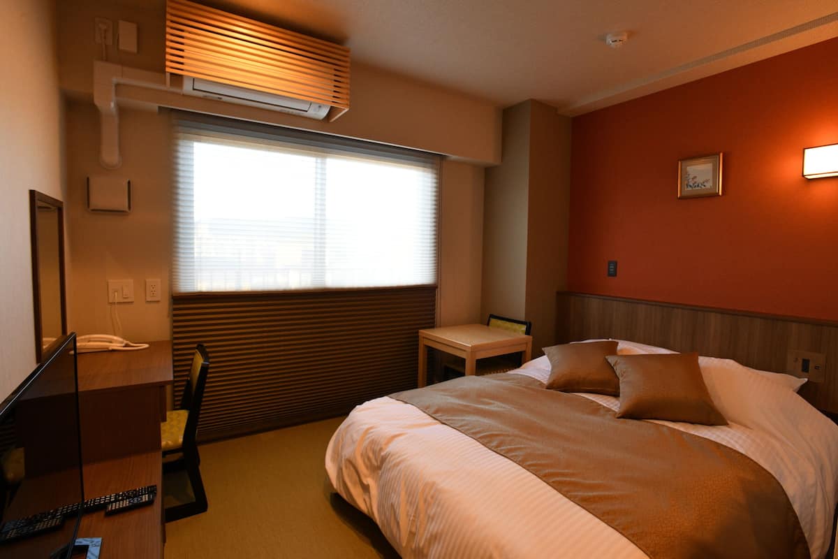 A hotel room with a bed, a desk with a telephone, a chair, and a window with blinds.