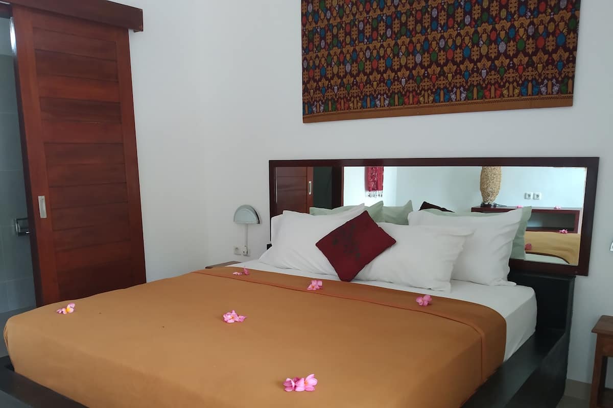 A hotel room with a bed, a wooden door, a wall-mounted mirror, and a decorative wall hanging.