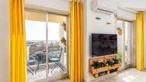 Una sala de estar moderna con un televisor grande, un banco de madera con plantas y un balcón con mesa y sillas.