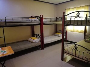 A dormitory room with bunk beds, a small table with items, and a window with curtains.