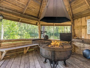 A rustic wooden cabin with a large outdoor cooking area, a bench, and a view of a forested area.