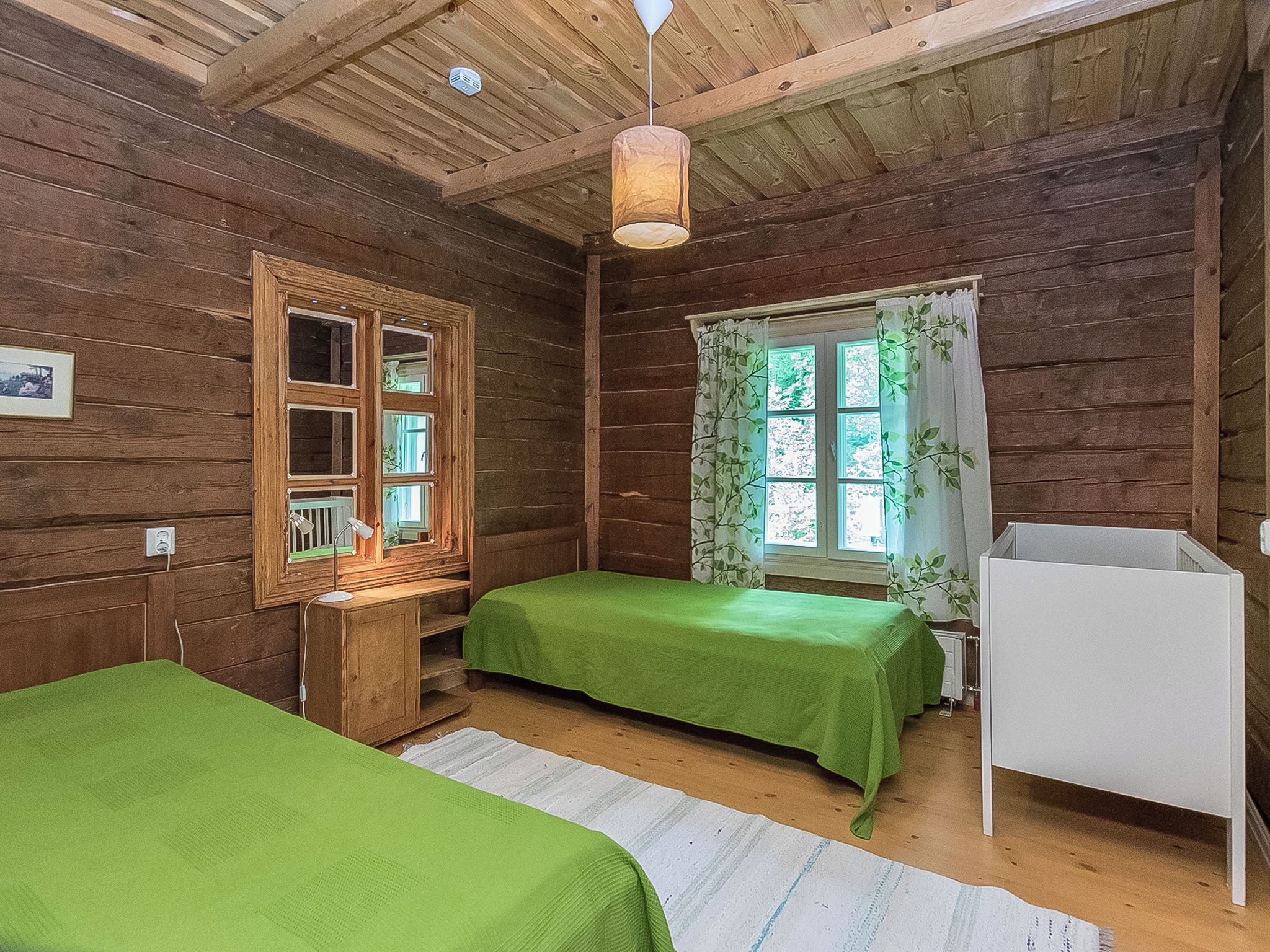 A wooden cabin room with two beds, a window with curtains, a desk with a lamp, and a white cabinet.