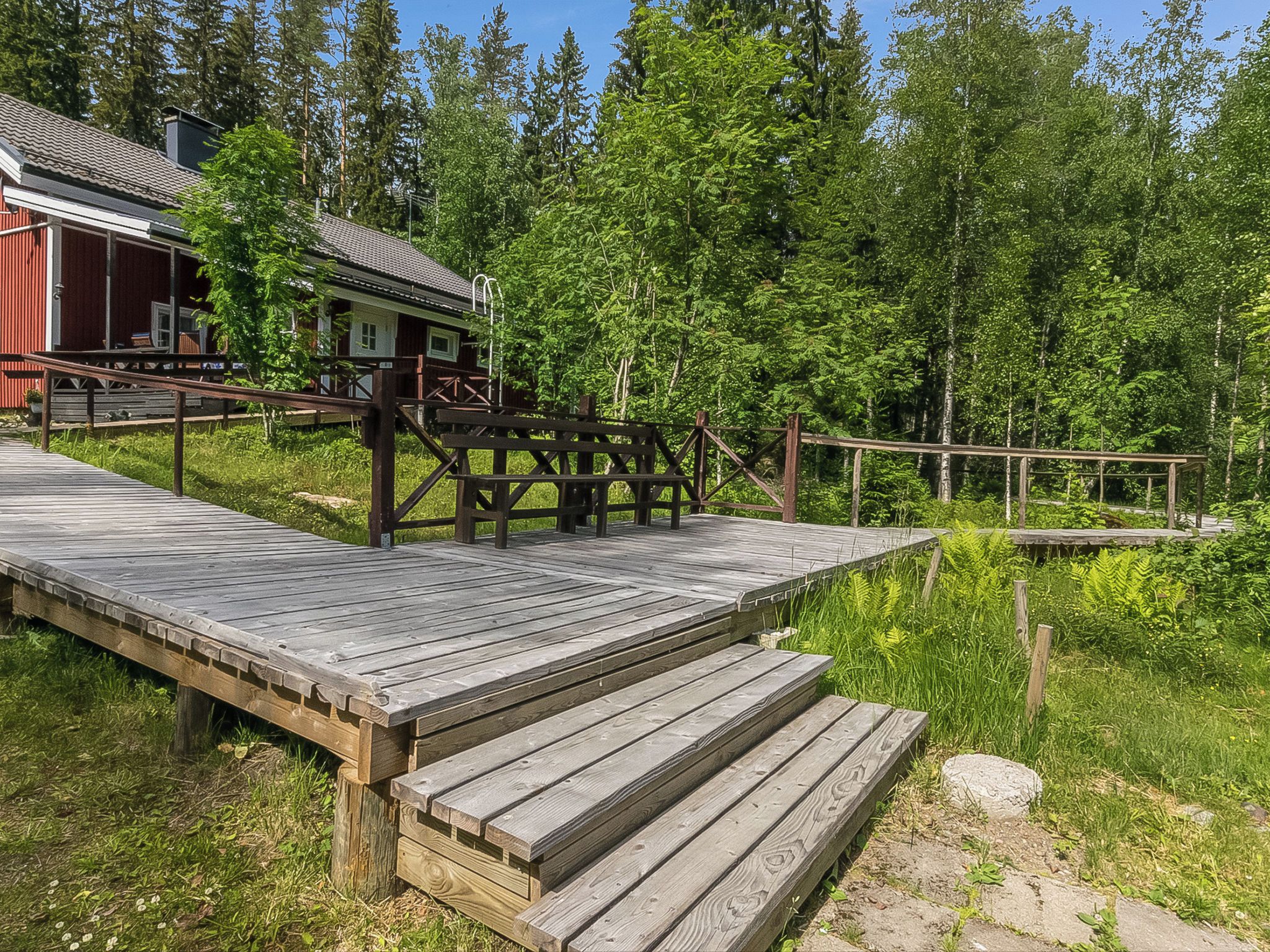 Puinen terassi johtaa mökkiin, jossa on metallinen kaide, ja terassin ympärillä on tiheä metsä.