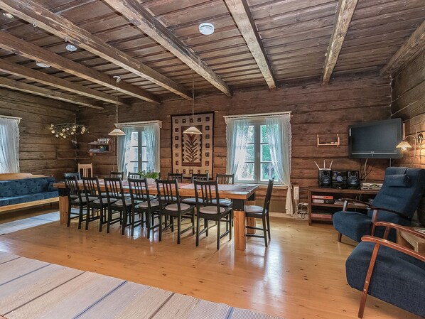 A rustic cabin interior with a wooden dining table surrounded by chairs, a sofa, and a television on a wooden stand.