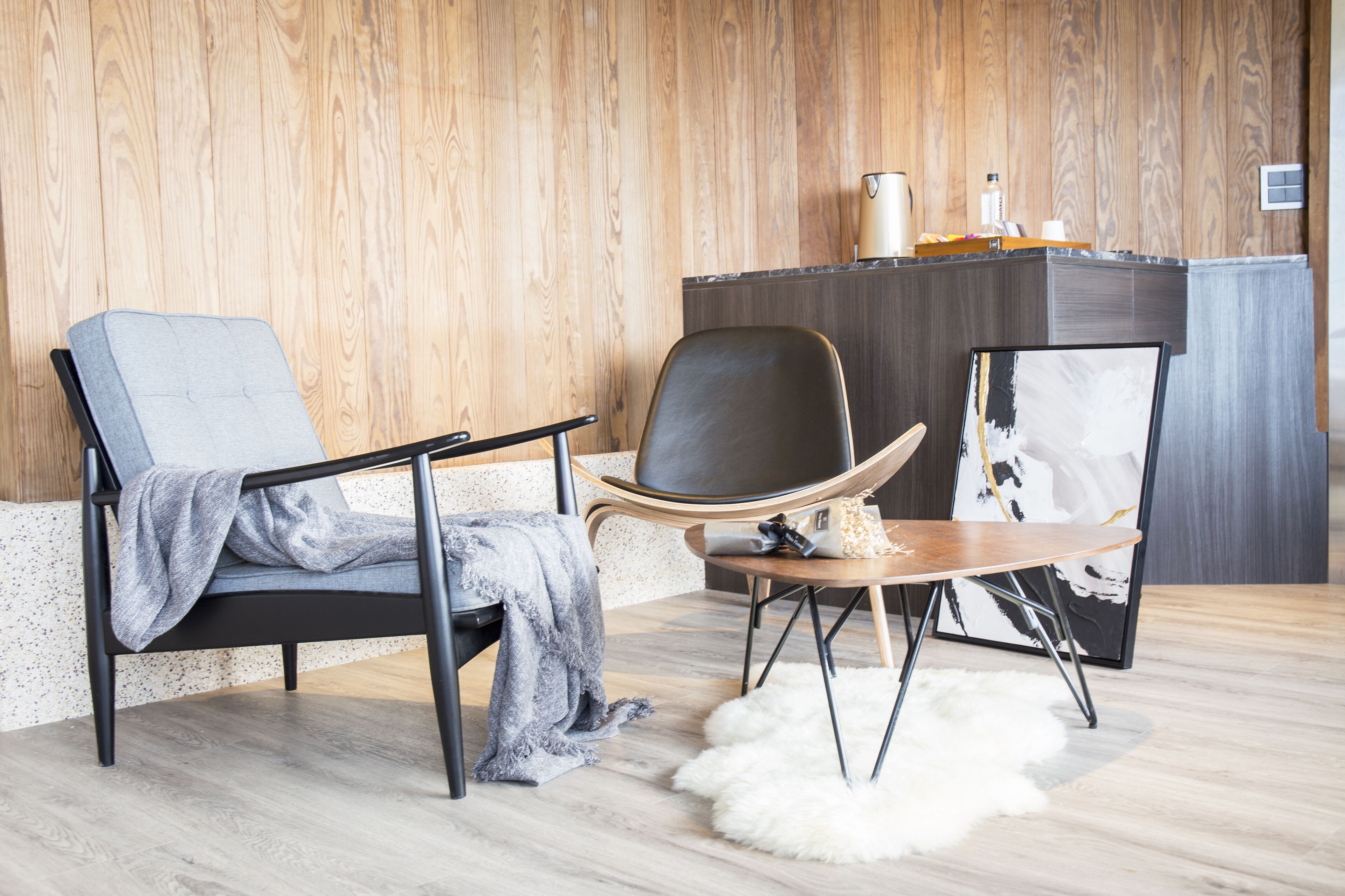 A modern living room with two chairs, a small table, a white furry rug, and a wooden wall.