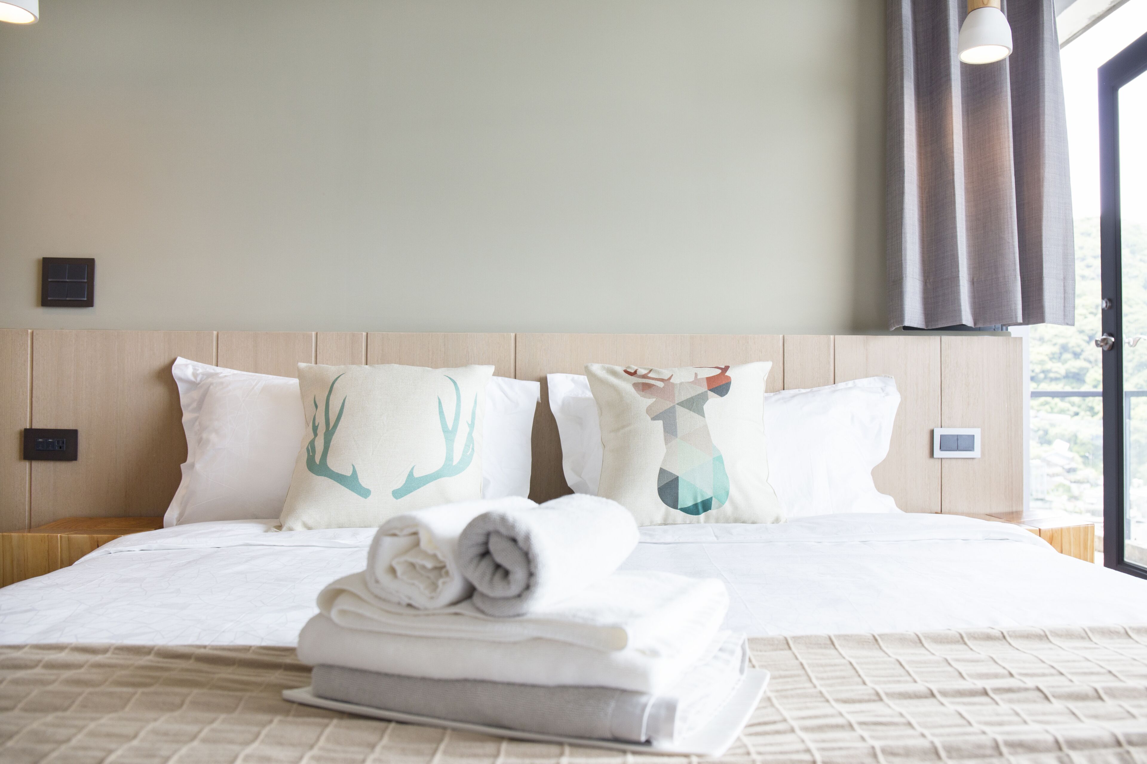 A neatly made bed with white linens and rolled towels, a wooden headboard, and a view of the outdoors through a window with curtains.