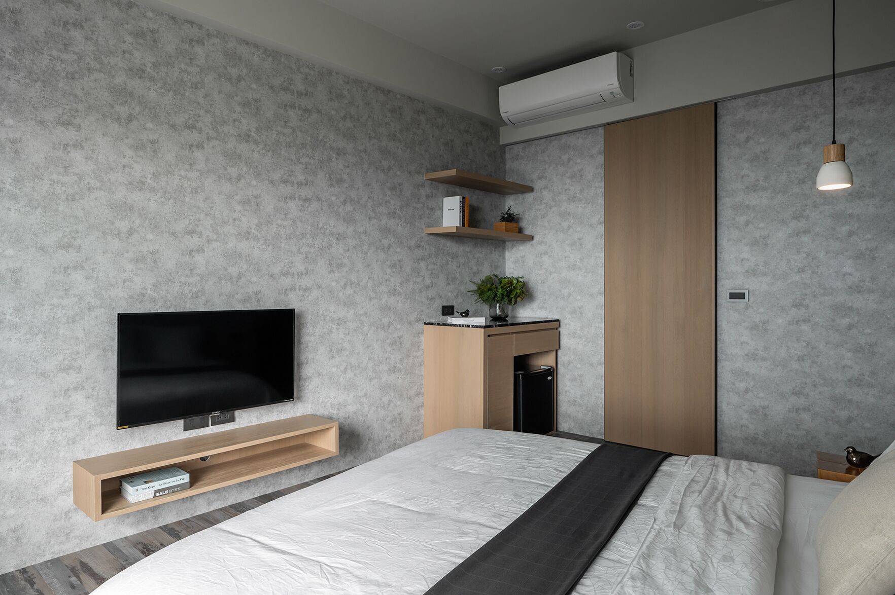 A modern bedroom with a flat-screen TV, a bed with a white and gray bedspread, a wooden nightstand, a small desk with a plant, and a wardrobe.