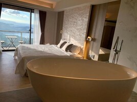 Un dormitorio moderno con una bañera grande, una cama con ropa de cama blanca y vistas al exterior.