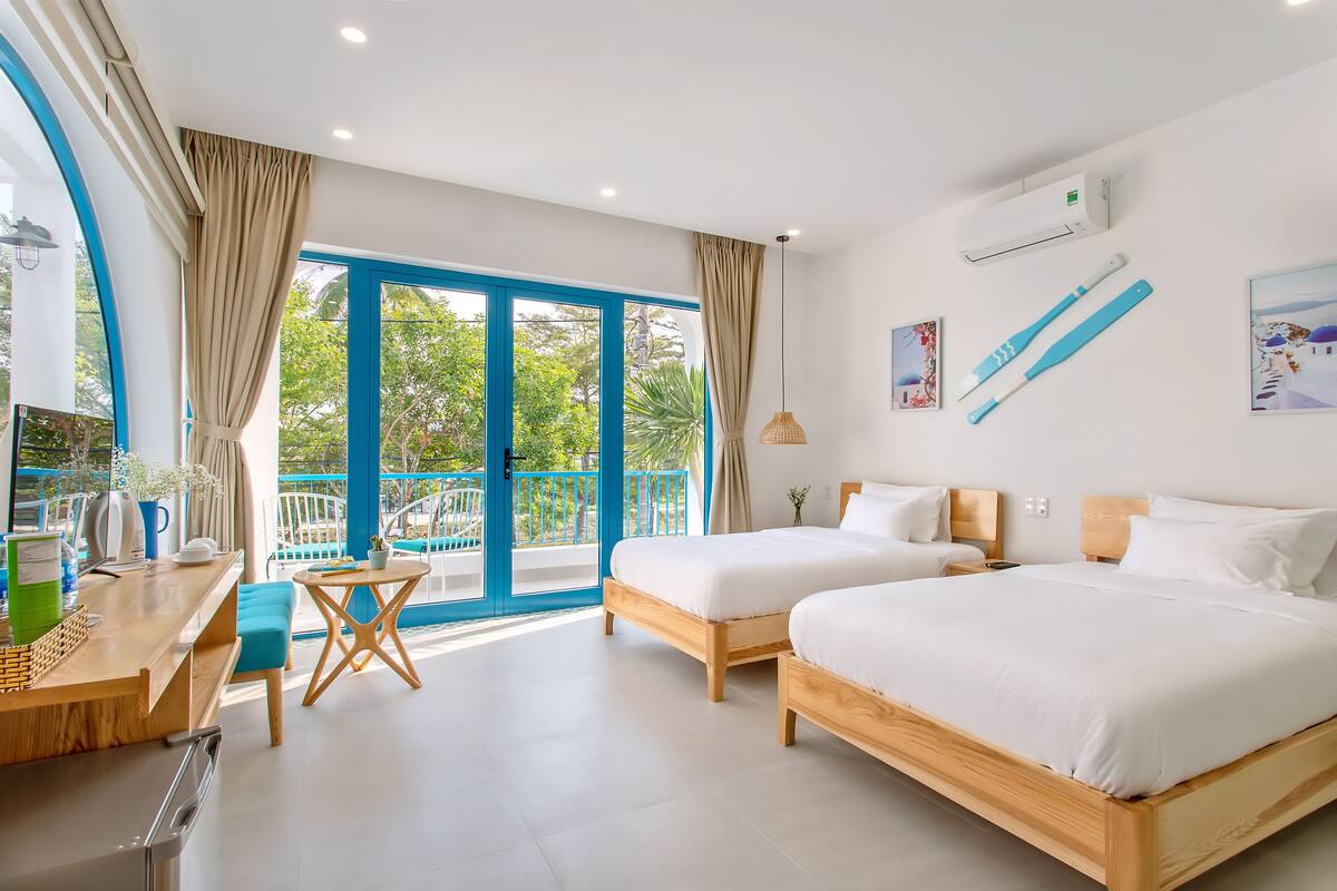 A modern hotel room with two beds, a wooden desk, a small table, and a balcony with blue doors.