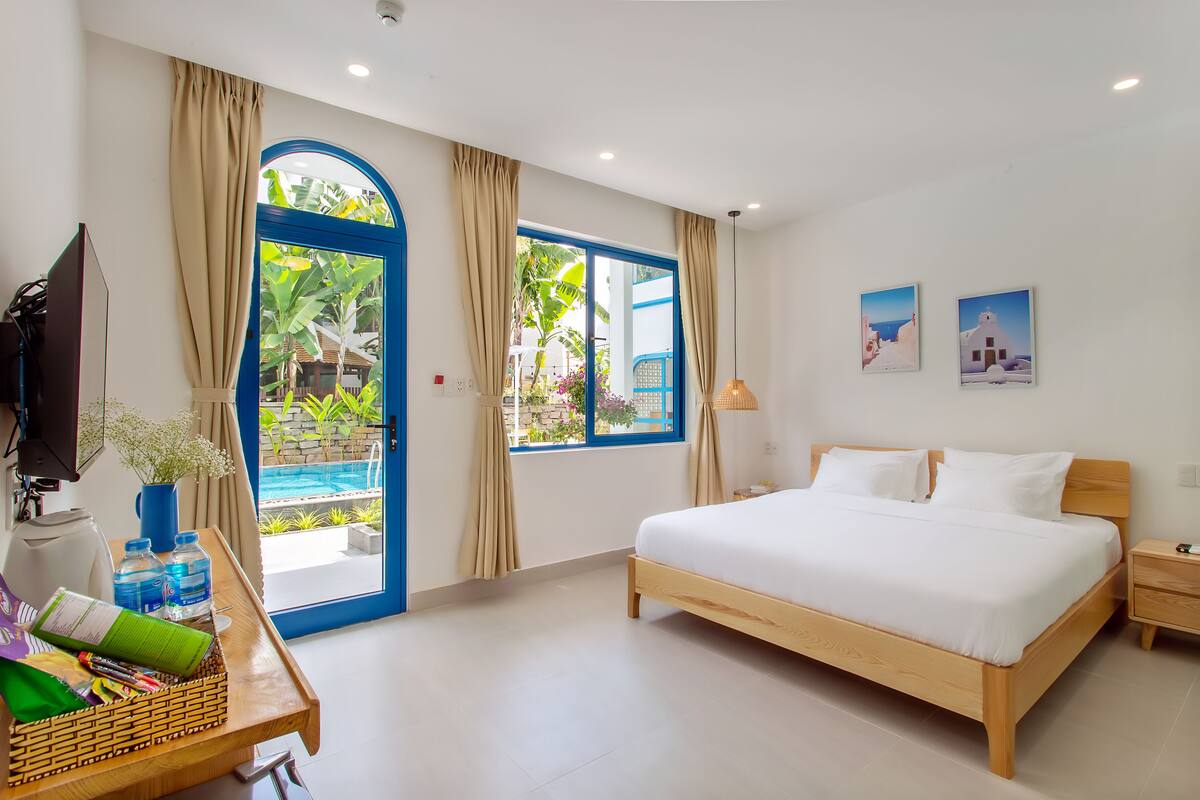 A hotel room with a bed, a desk with a TV, a window with curtains, and a view of a pool and greenery.