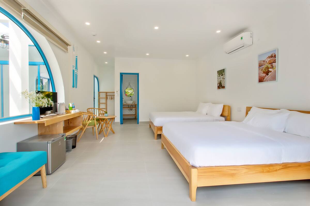 A modern hotel room with two beds, a desk, a small refrigerator, and a large window with blue trim.
