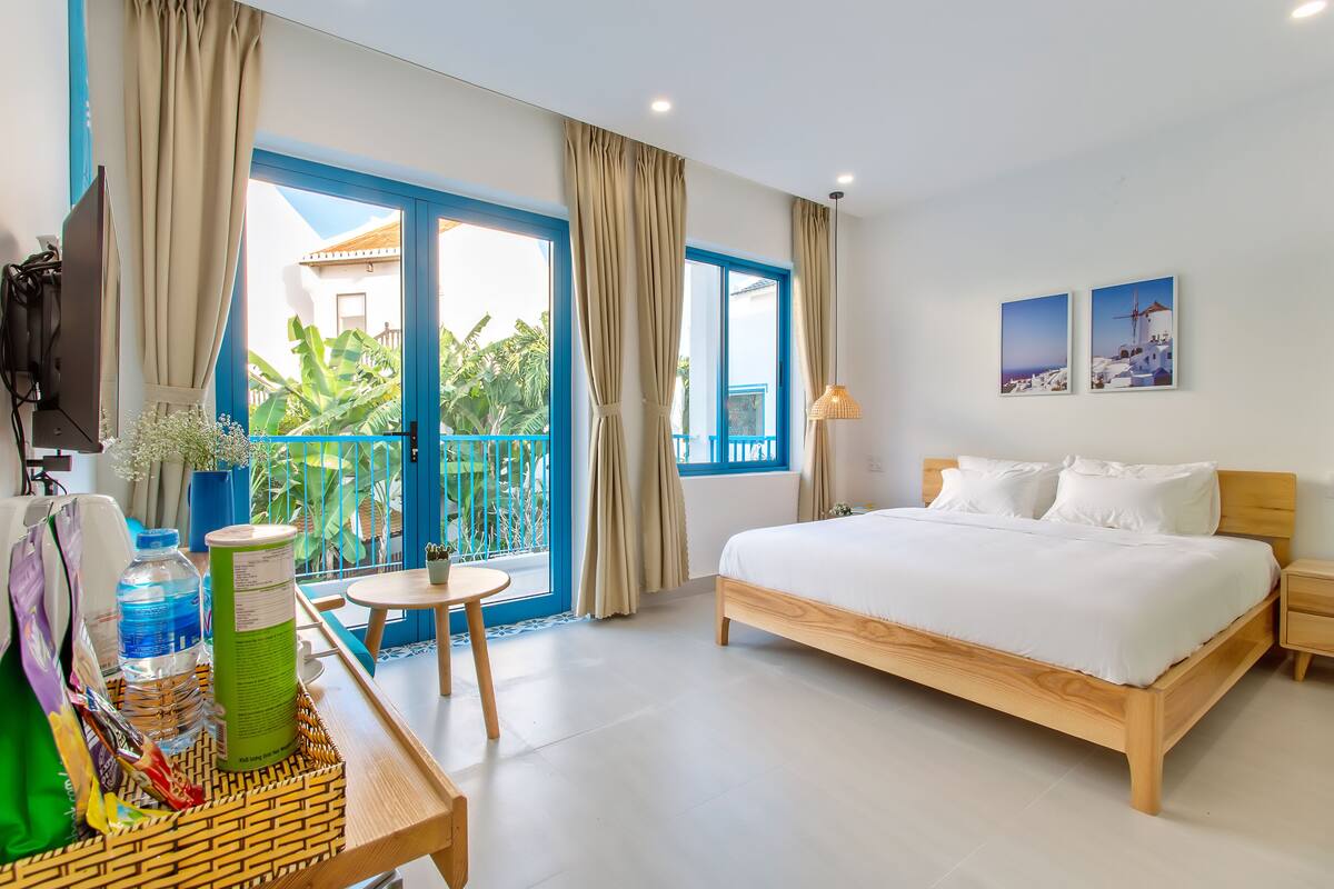 A hotel room with a bed, a small table with a bottle and a bag, a TV, and a balcony with curtains.