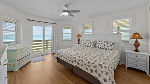 Un dormitorio con una cama, un ventilador de techo, una mesita de noche con lámpara y vista al océano a través de las ventanas.