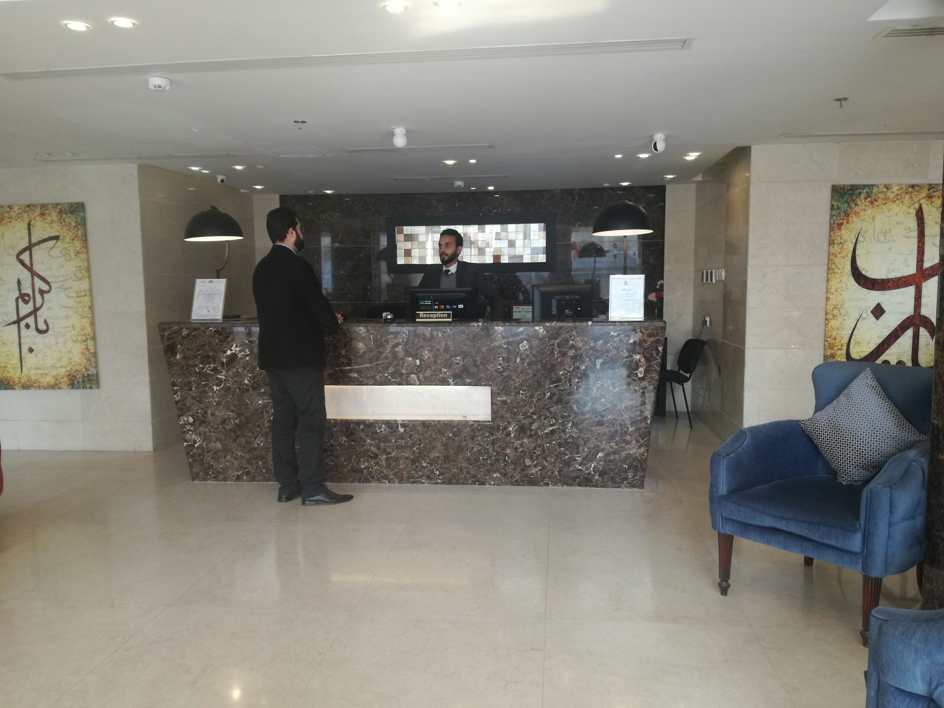 A reception desk with a marble top, a man standing in front, and a blue armchair with a patterned pillow to the right.