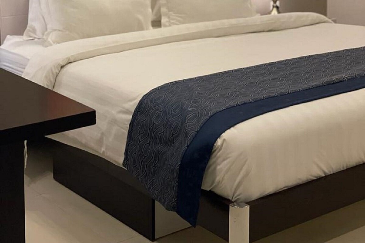 A bed with white linens and a blue blanket, a bedside lamp, and a colorful abstract painting on the wall.