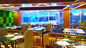 A restaurant with multiple dining tables and chairs, a green wall, and a view of the cityscape through large windows. - Hotel Grand View (Sylhet)